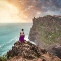 Man standing on cliff with scenic view of Uluwatu Temple, Bali at sunset.